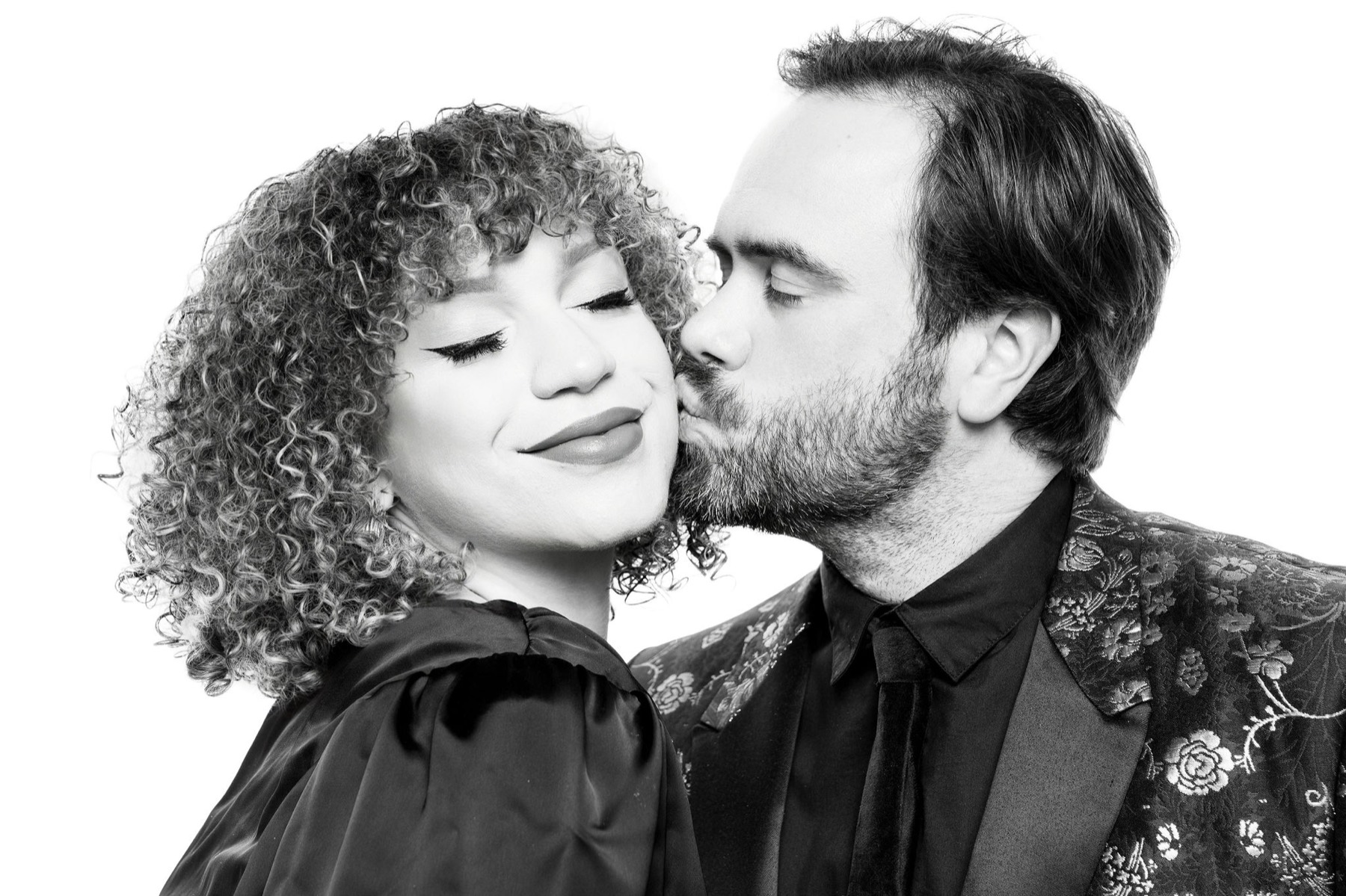 A man kissing his partner's cheek as she smiles, photographed against the white backdrop